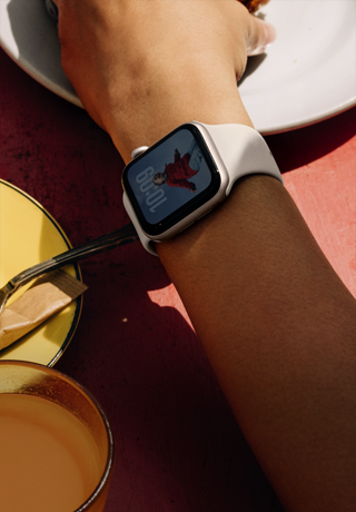 Arm van iemand die iets van een ontbijtbord pakt, Apple Watch SE 3 om de pols, kast van aluminium, kleur Sterrenlicht, Always-On display, portret van een persoon op de wijzerplaat, sportbandje, kleur Sterrenlicht